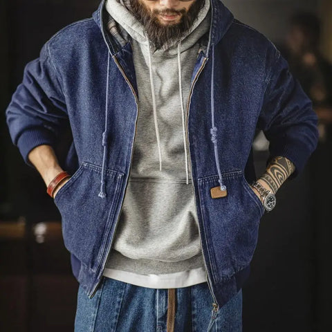 Man wearing a blue denim jacket over a gray hoodie with tattoos on arms.