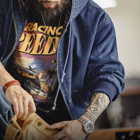 Person wearing a denim jacket and graphic t-shirt with a tattooed arm, holding a brown satchel.